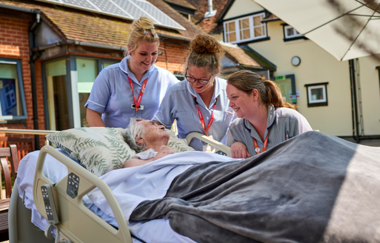 3 nurses smiling standing next to a patient in bed in the garden