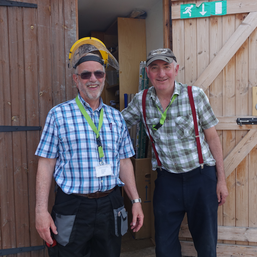Two male maintenance volunteers smiling outside a maintenance shed